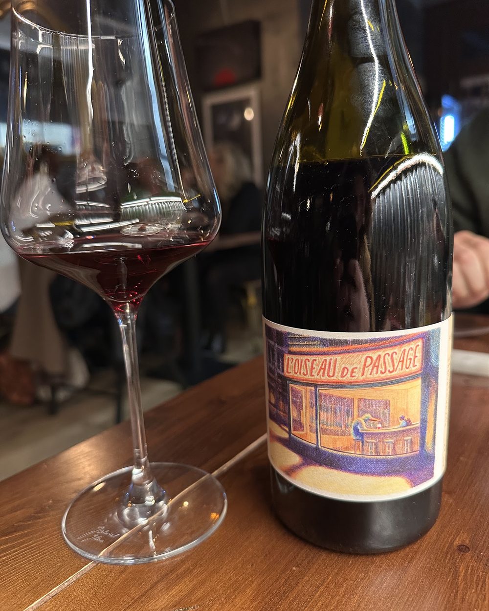 Un calice di vino rosso accanto alla bottiglia da cui &egrave; stato versato, su un tavolo di legno, con la sala illuminata alle spalle.