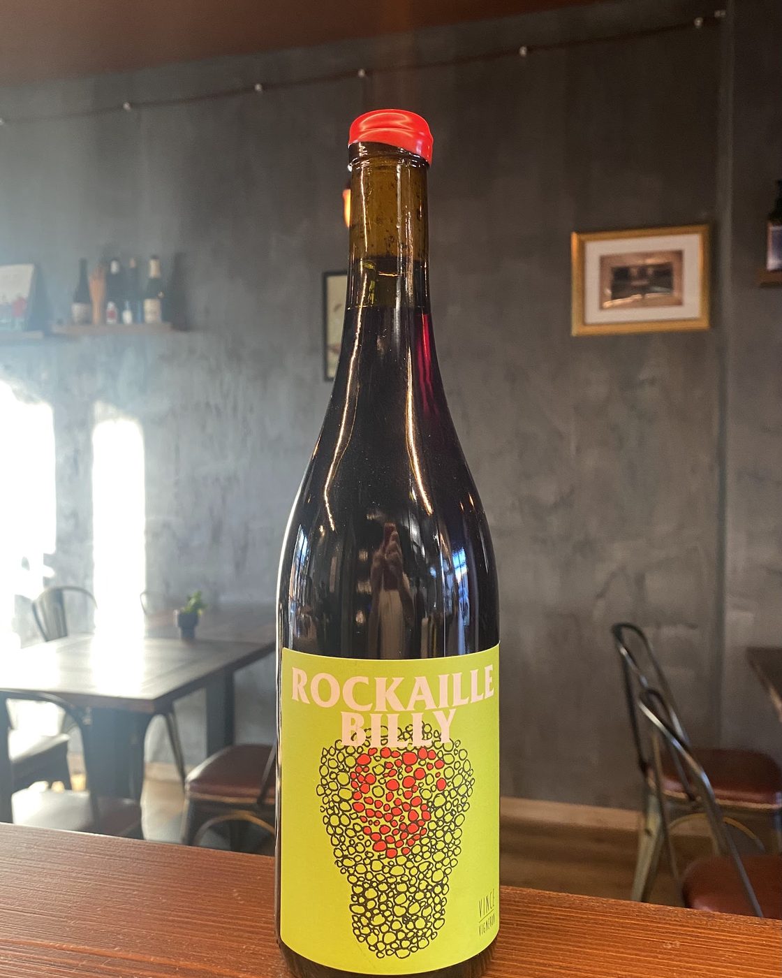 Una bottiglia di vino rosso artigianale aperta sul bancone di legno; dietro, la sala con pareti grigie smaltate, sedie da bistrot in ferro e una mensola di bottiglie.